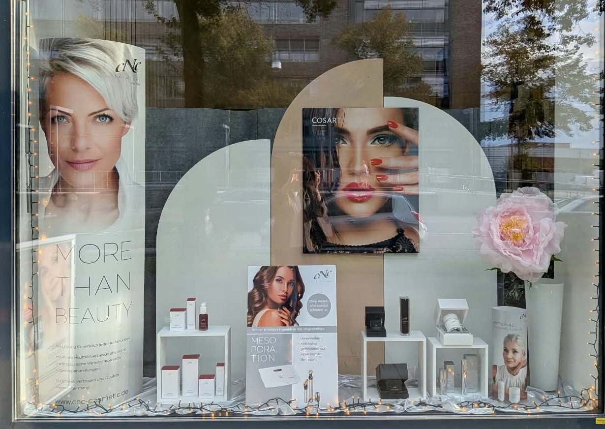 Zweites Schaufenster mit zwei hellen Rundbögen, Pflegeprodukten auf weißen Podesten und einer rosa Pfingstrose in einer weißen Vase