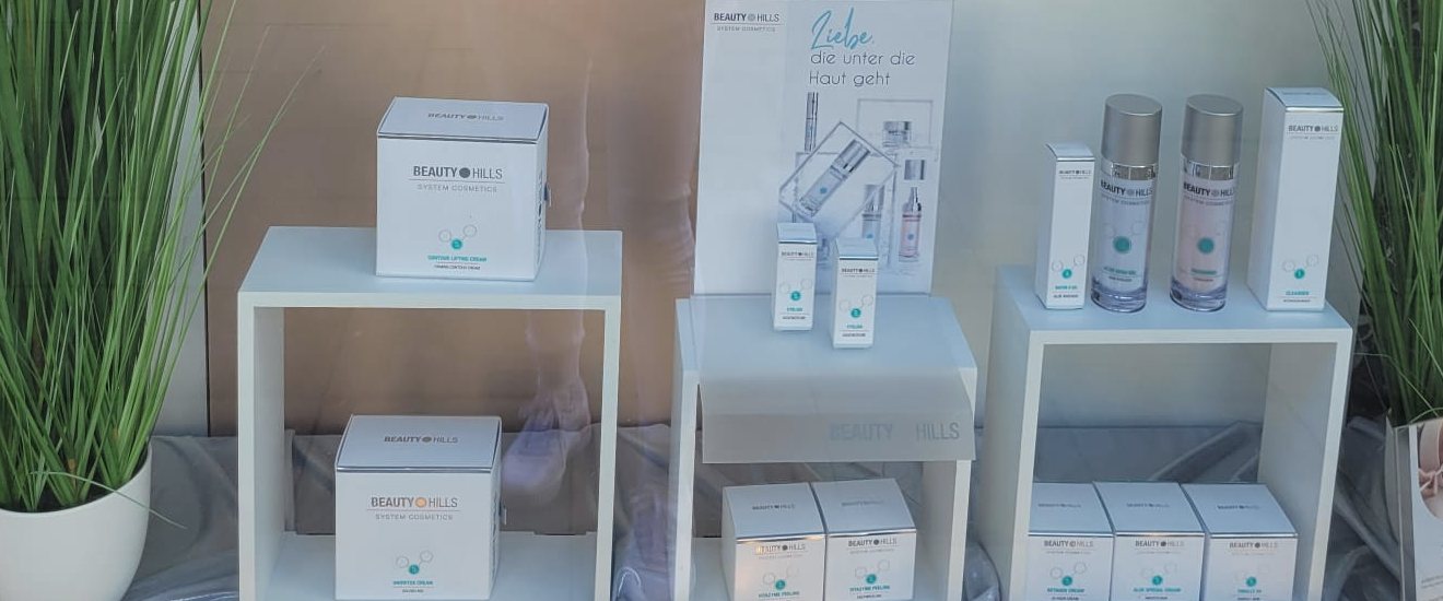 Pflegeprodukte von Beauty Hills auf weißen Podesten im Schaufenster, zwischen zwei Ziergräsern